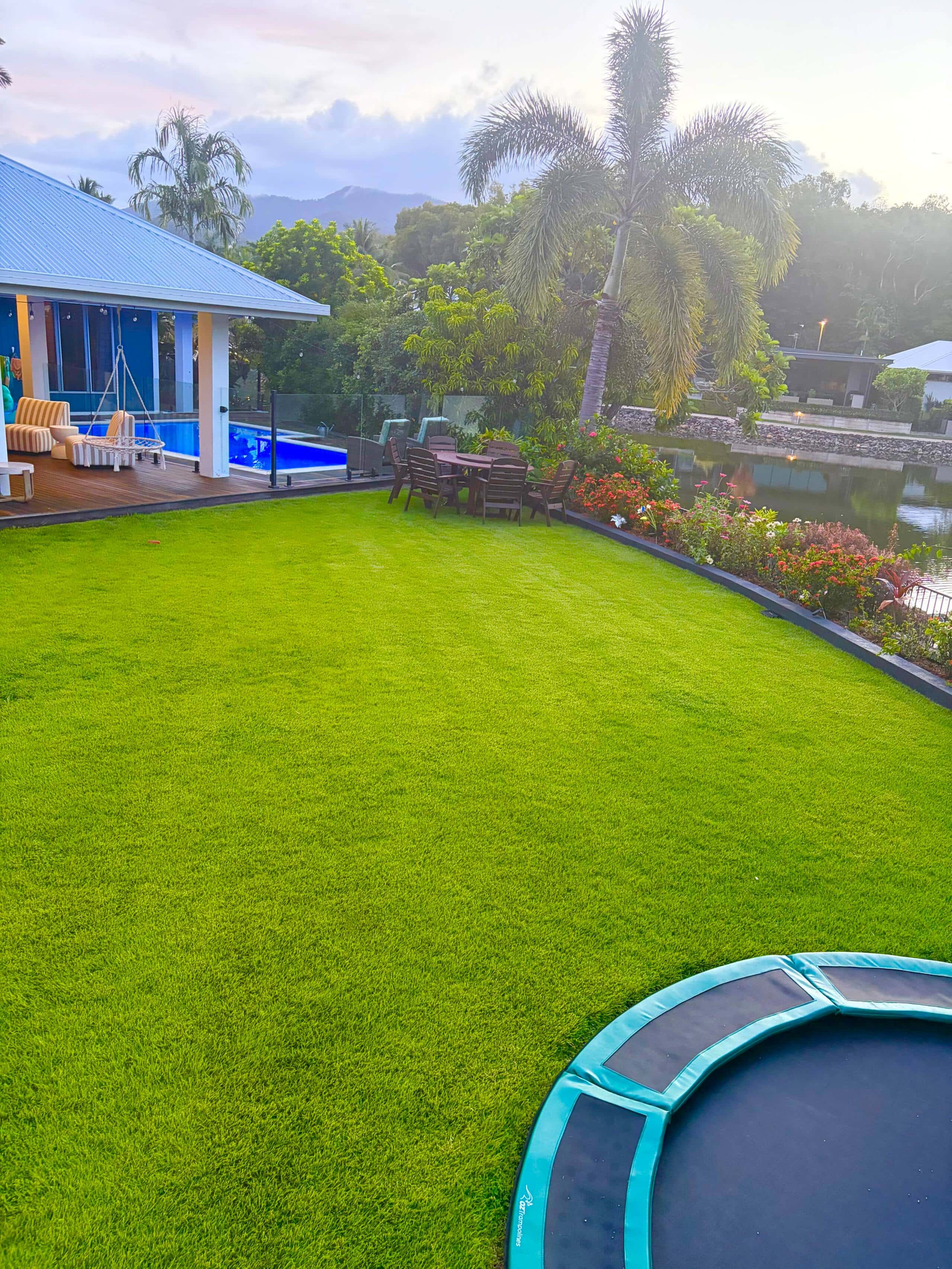Fully fenced backyard lawn with in-ground trampoline, pool deck and lagoon at sunrise
