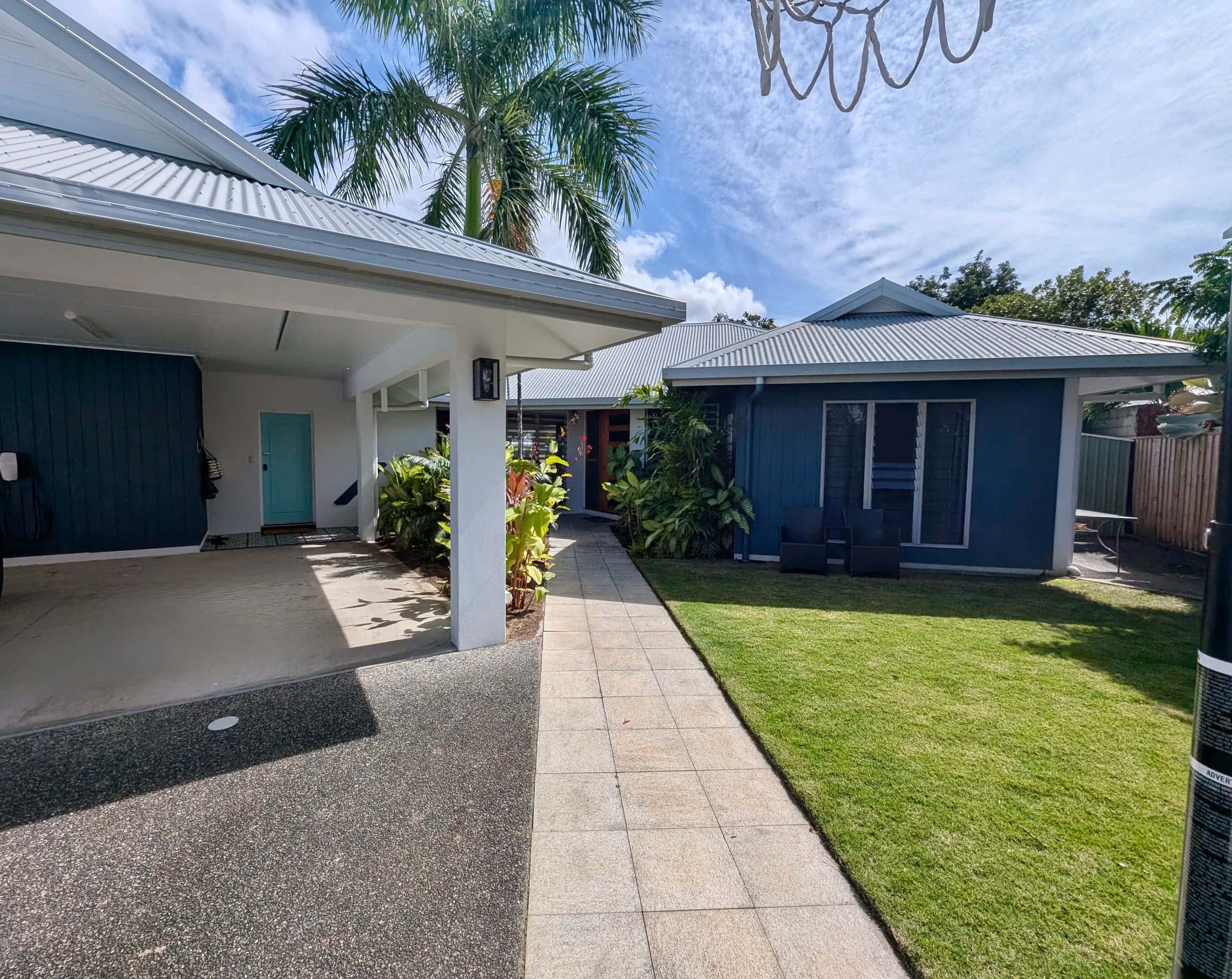 Front of the house with covered carport, aqua front door, blue weatherboard and tropical gardens