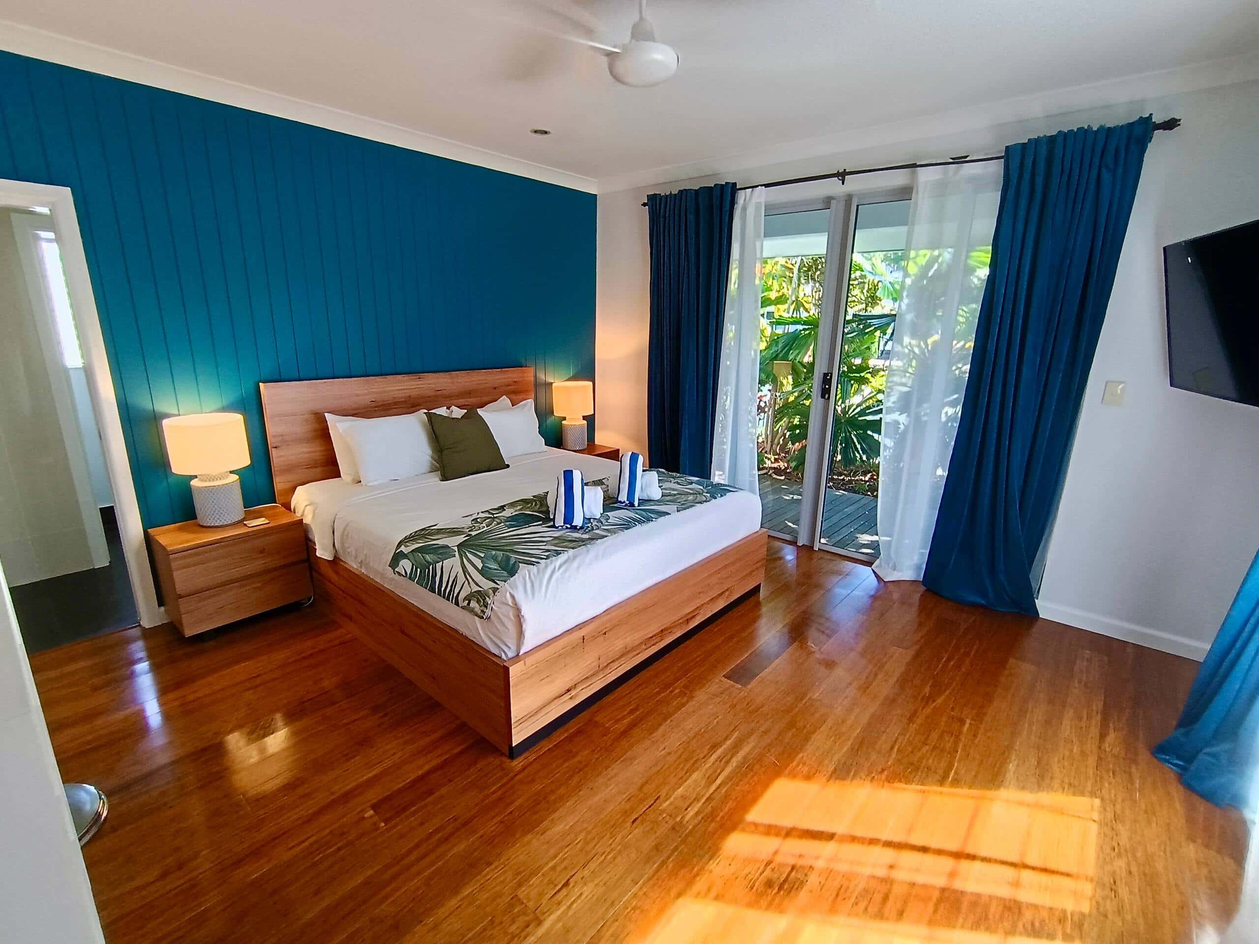Master bedroom with king bed on a gas-lift storage base, timber bedhead, teal panelled wall and a sliding door out to the pool