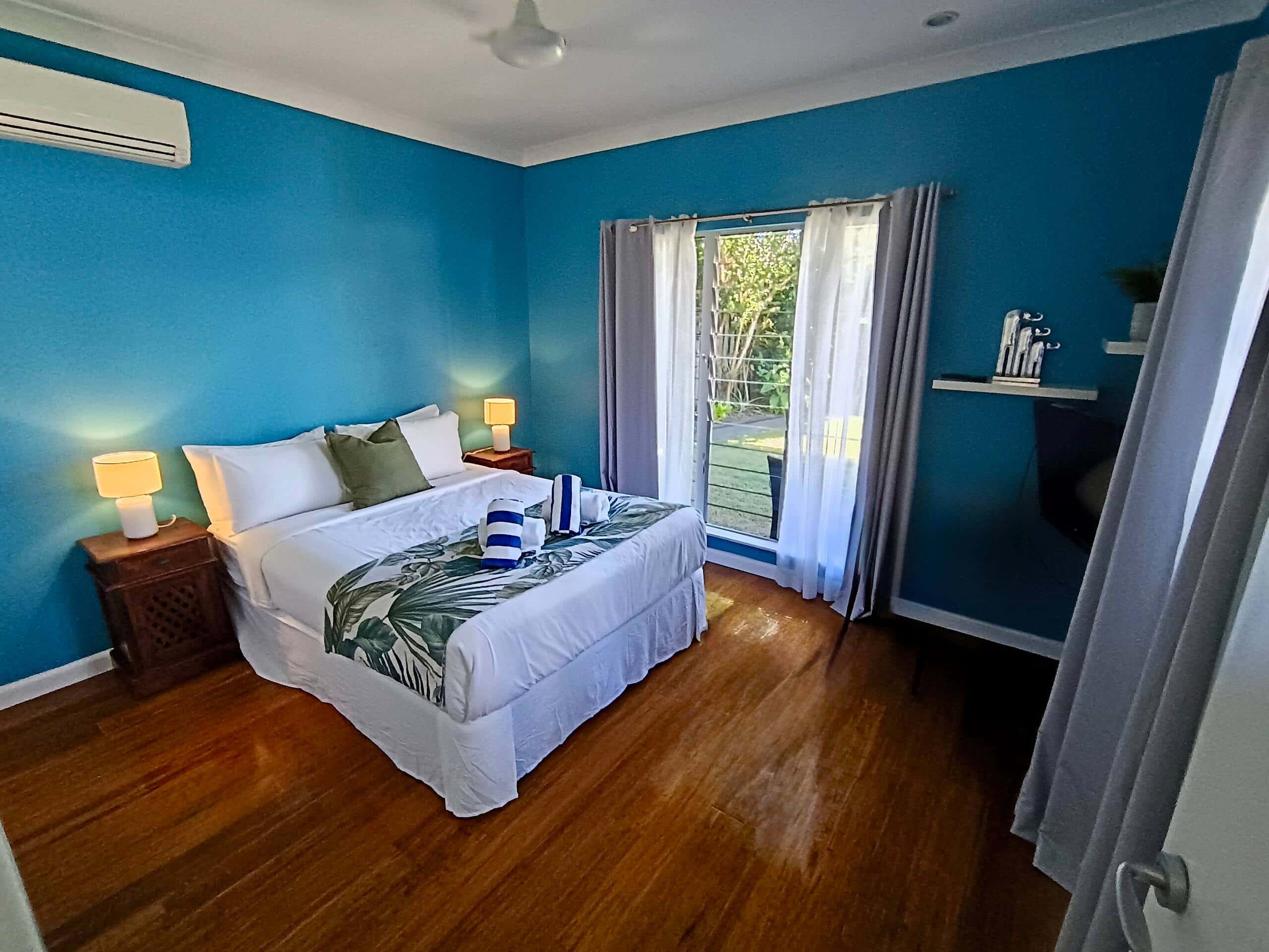 Queen bedroom with teal feature walls, wall-mounted TV and louvre windows looking onto the garden